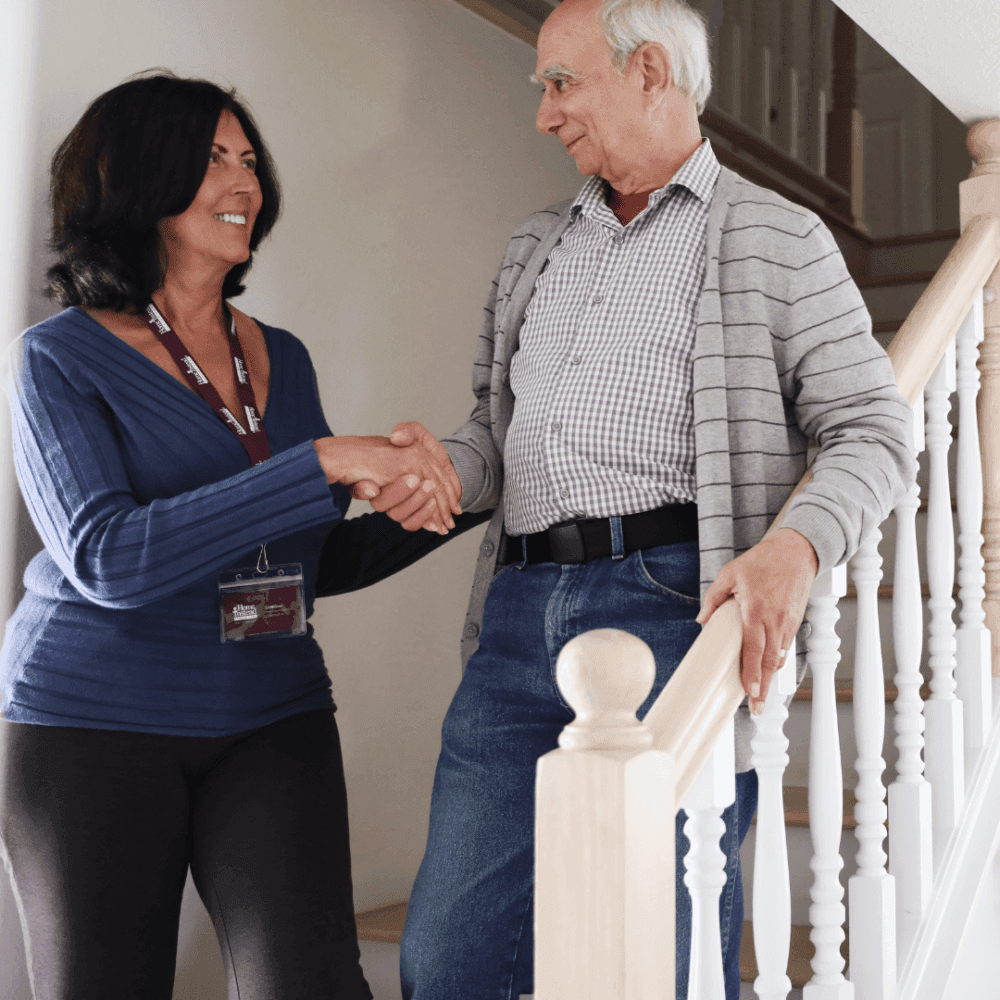 A woman and an elderly man shaking hands at the bottom of a staircase, both smiling. - Home Instead