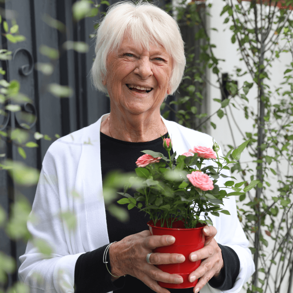 Elderly woman smiling and holding a red pot with pink flowers, surrounded by greenery. - Home Instead
