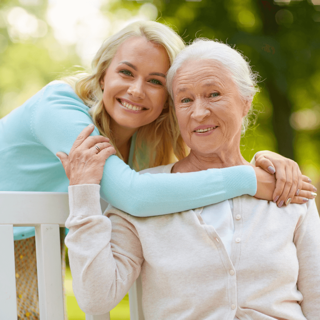 A smiling young woman embraces an elderly woman on a park bench, both looking at the camera. - Home Instead