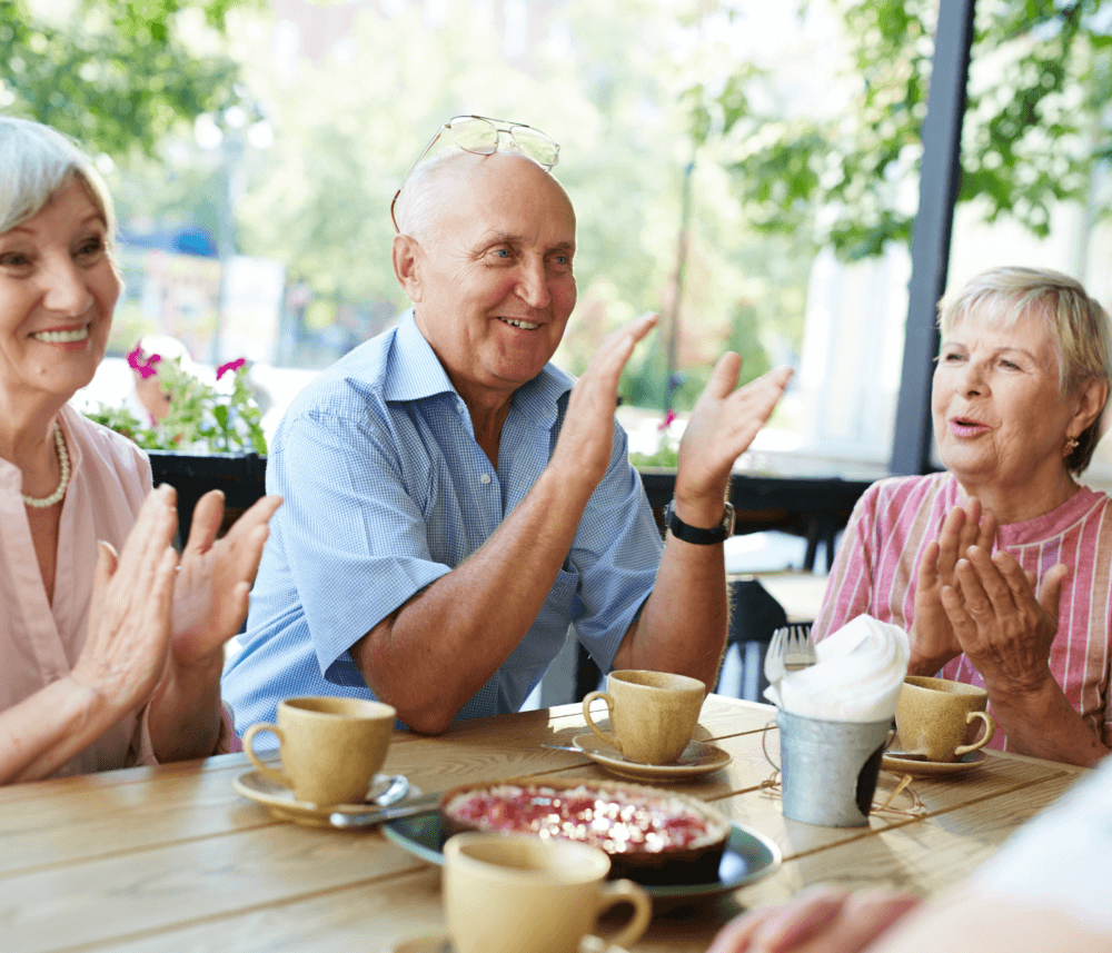 Three elderly people applauding while sitting at a table with coffee and a dessert in an outdoor café. - Home Instead