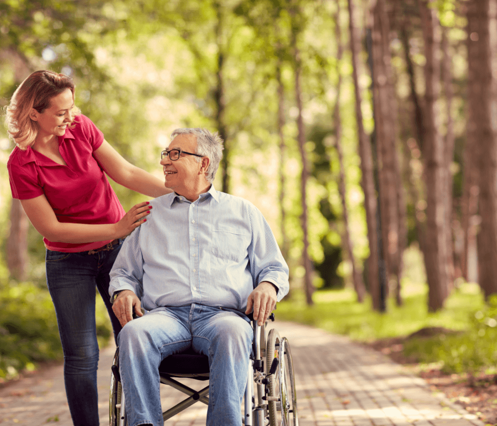 Woman in a red shirt smiling and pushing a man in a wheelchair along a tree-lined path on a sunny day. - Home Instead