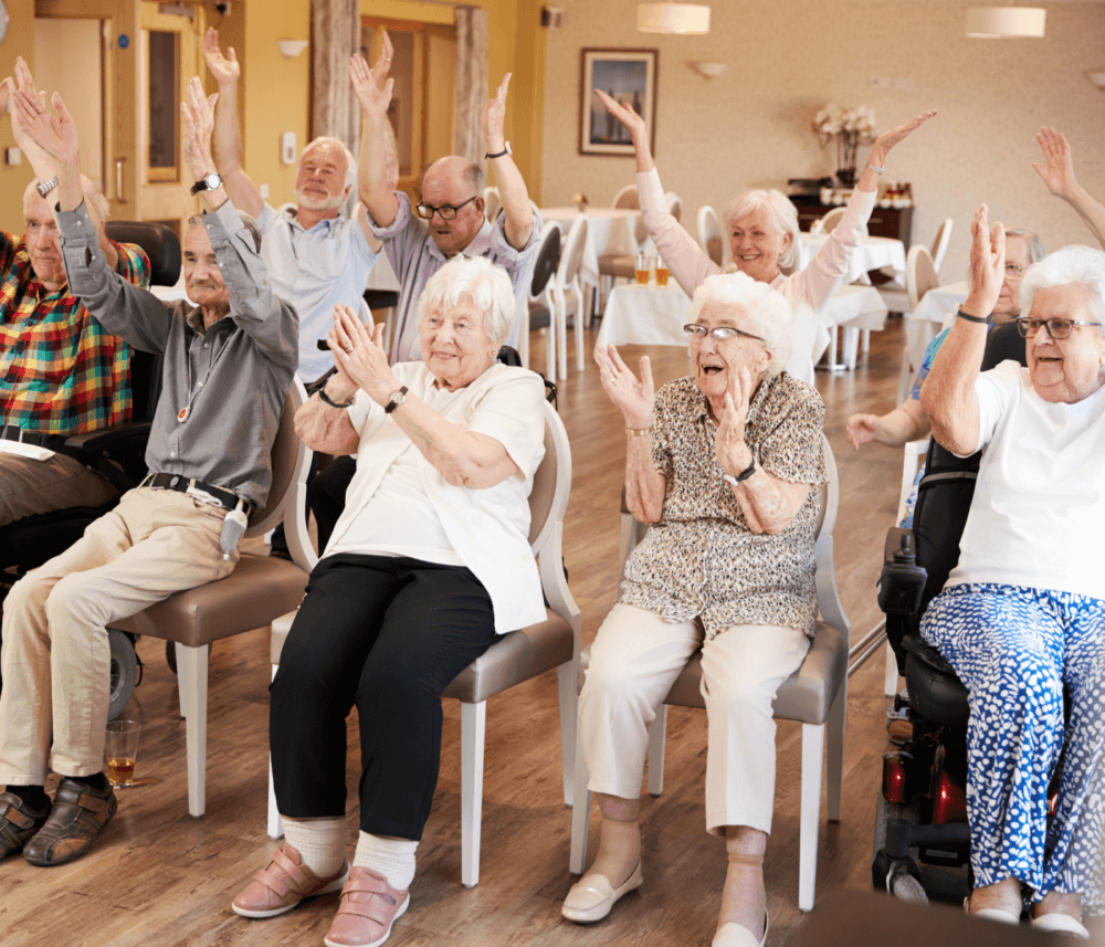 A group of seniors, some seated and some in wheelchairs, joyfully raising their arms and smiling in a communal room. - Home Instead