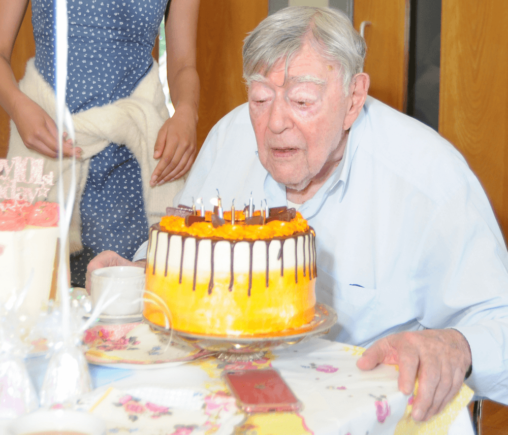 An elderly man leans forward to blow out candles on a decorated birthday cake while surrounded by people. - Home Instead