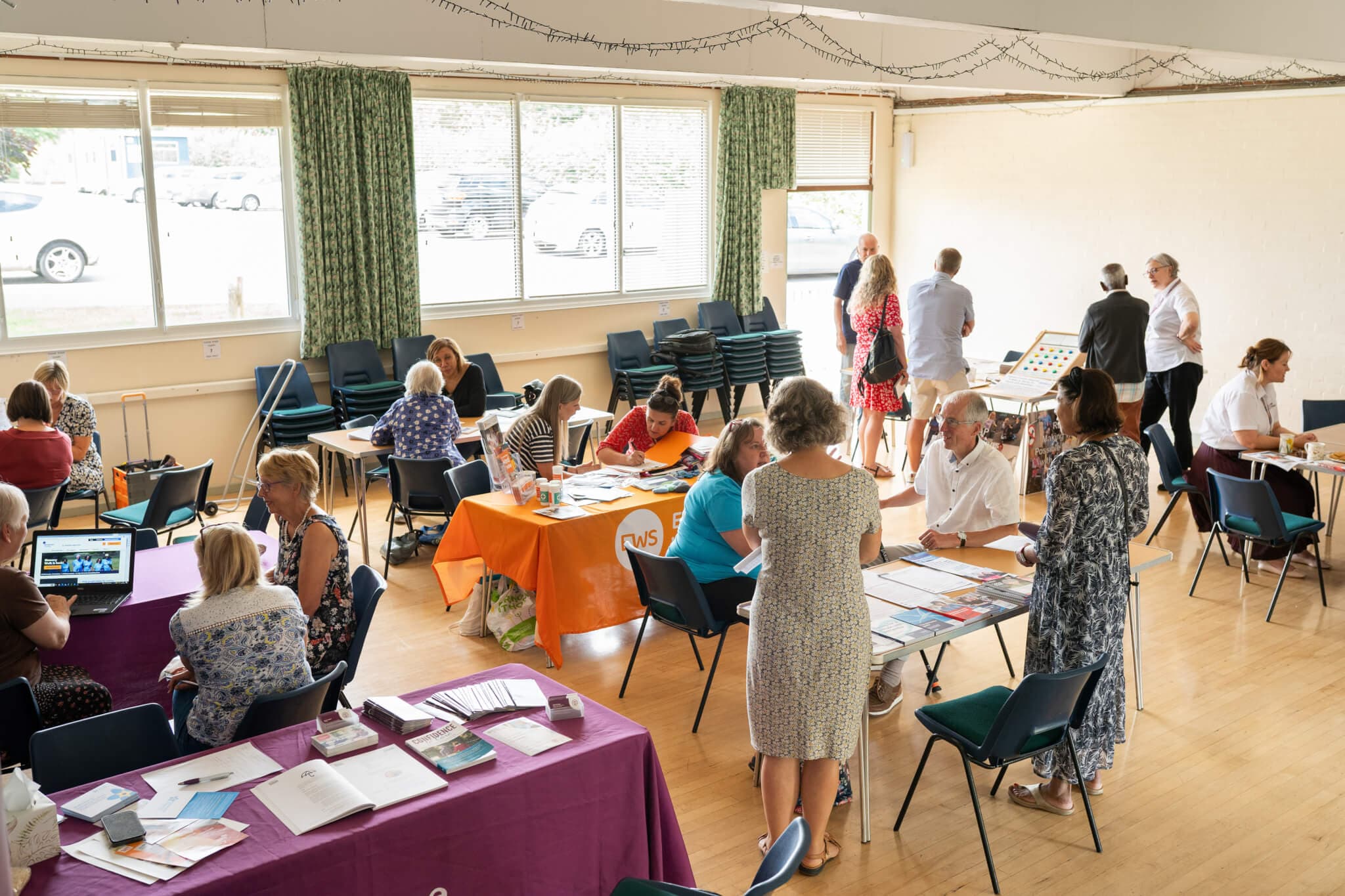 People attending a community event in a hall, sitting at tables with informational displays and talking with each other. - Home Instead