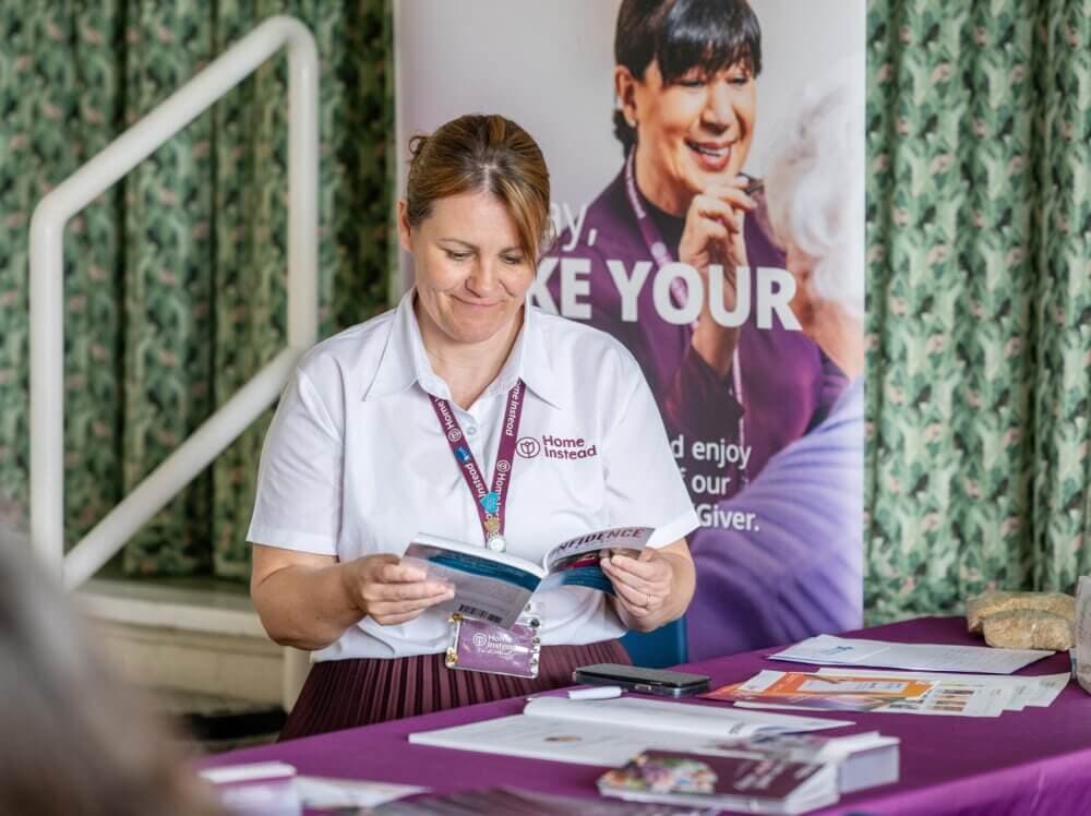 A woman in a white shirt reads a brochure at a table covered with papers and brochures. A banner stands behind her. - Home Instead