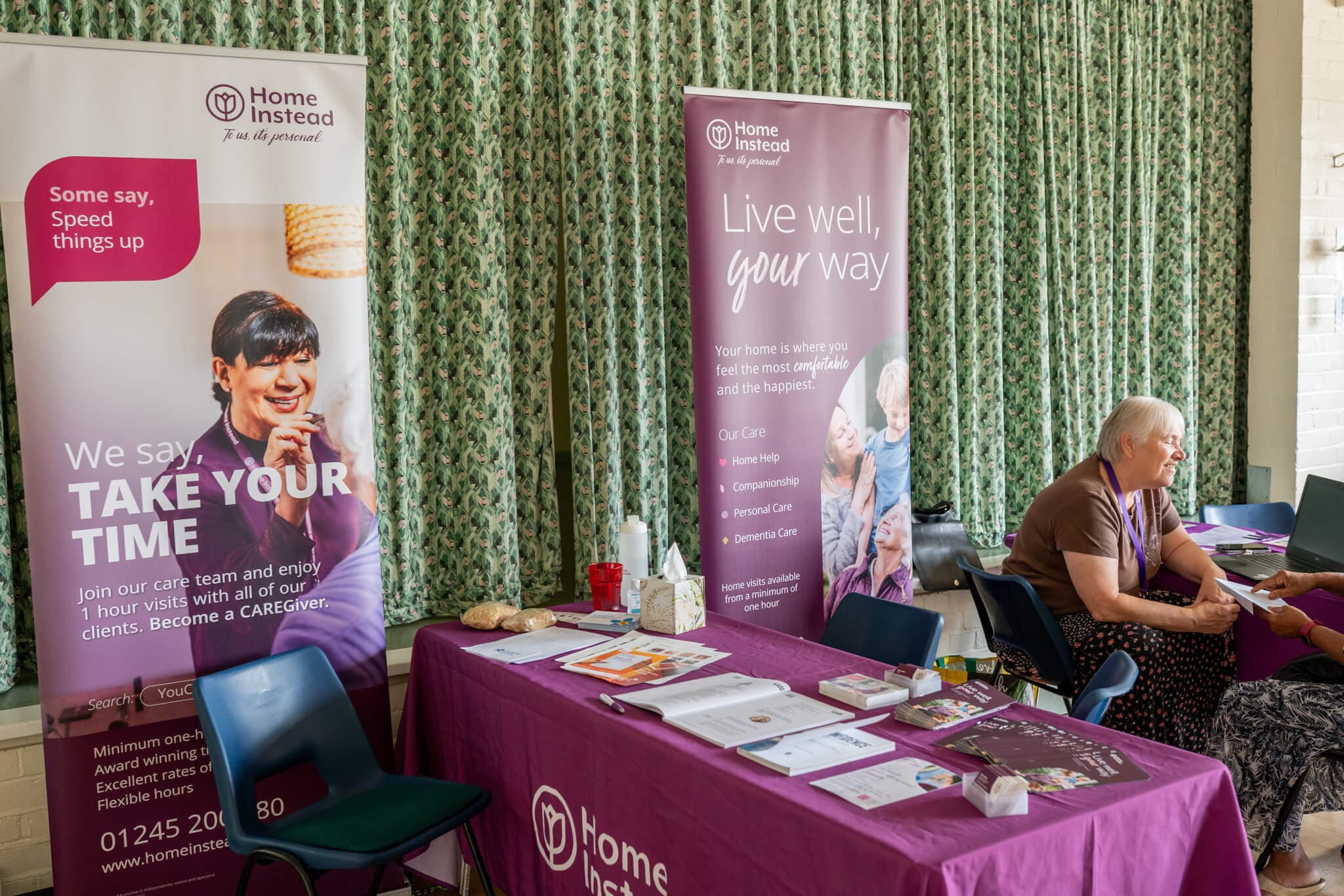 A booth with "Home Instead" banners and pamphlets. A person is sitting at the table covered with a purple tablecloth. - Home Instead