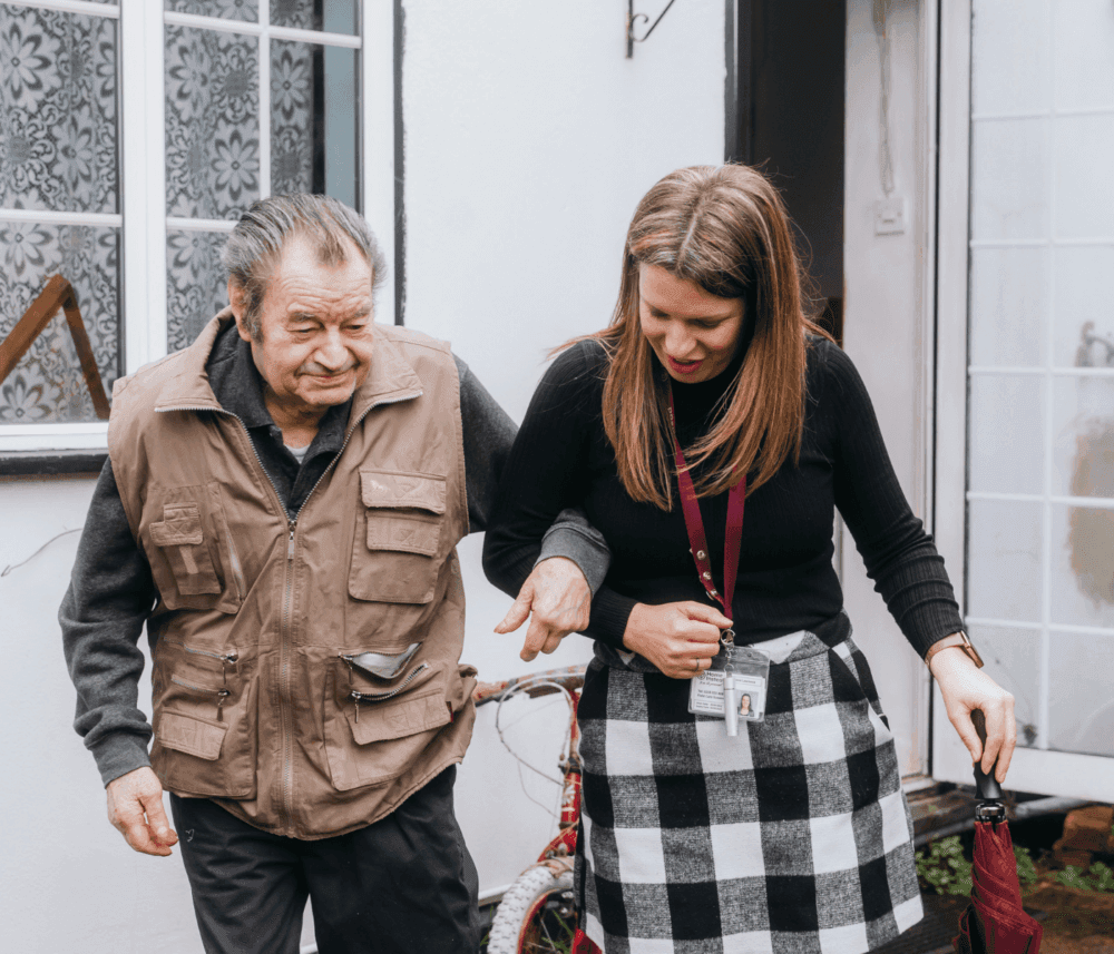An elderly man walks arm-in-arm with a woman, who appears to be assisting him, outside a house. - Home Instead