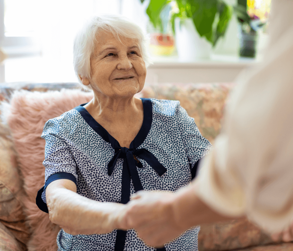 Elderly woman with white hair, smiling and holding hands with another person indoors. - Home Instead