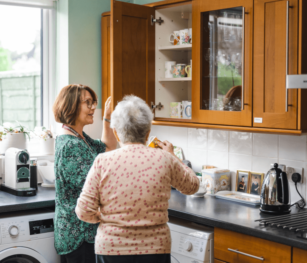 Two women in a kitchen, one opening a cabinet and the other holding a box while they talk and organize items. - Home Instead