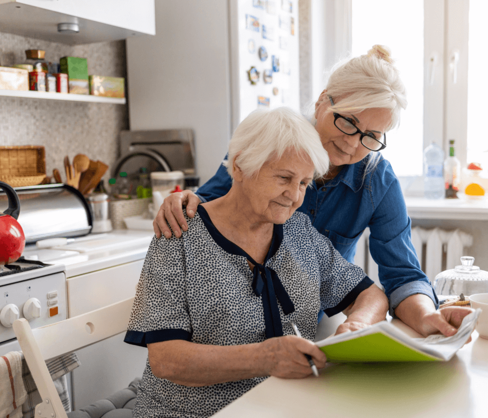 Older woman and elderly woman reviewing a document together at a kitchen table, both looking attentive. - Home Instead