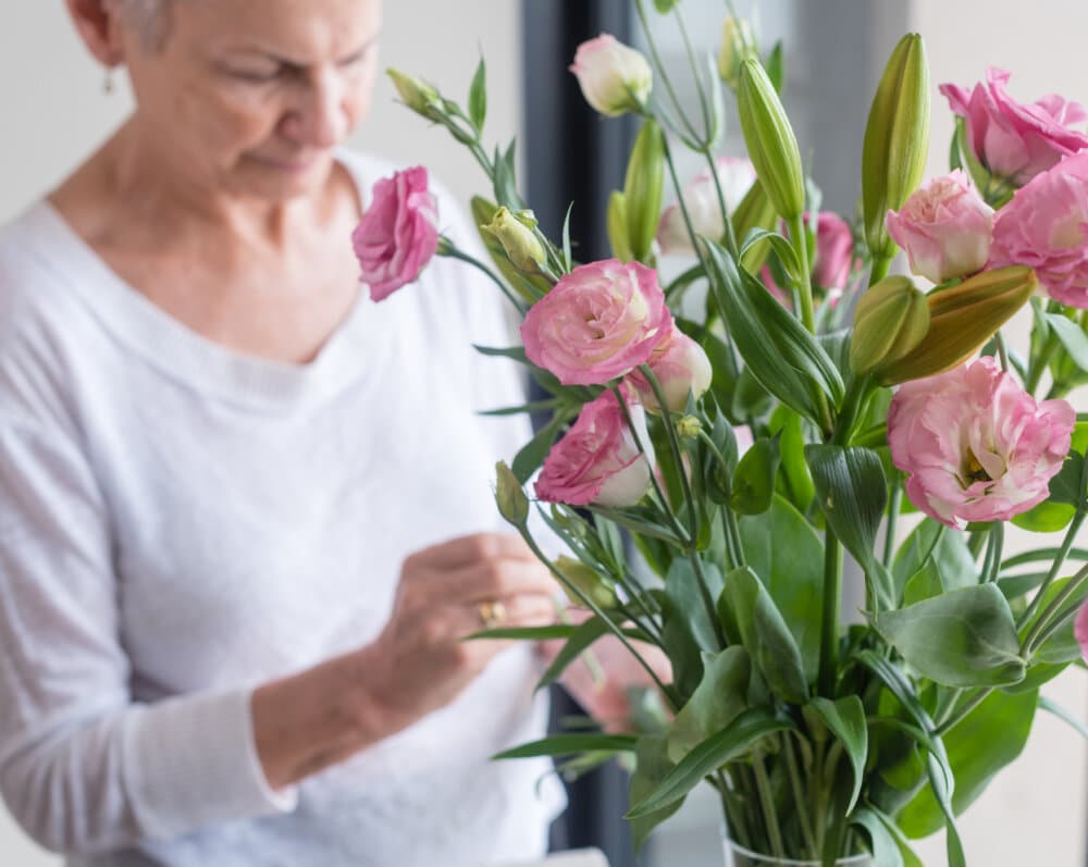 An elderly woman arranging a bouquet of pink and white flowers in a bright, indoor setting. - Home Instead