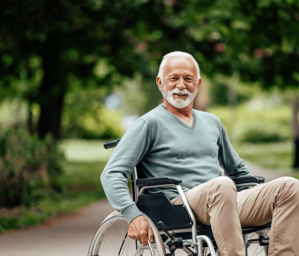 A smiling elderly man with a white beard in a wheelchair, wearing a light green sweater, sits outdoors in a park. - Home Instead