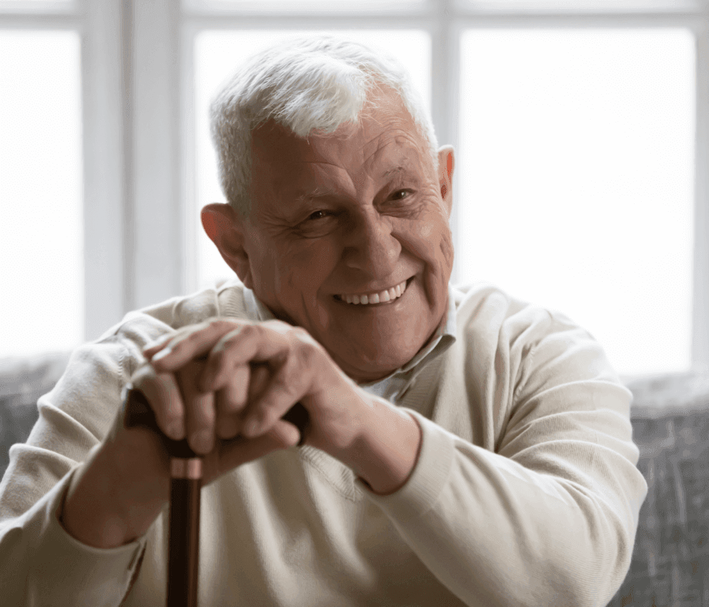 Smiling elderly man with white hair, wearing a beige sweater, resting on a cane, sitting against a bright window. - Home Instead