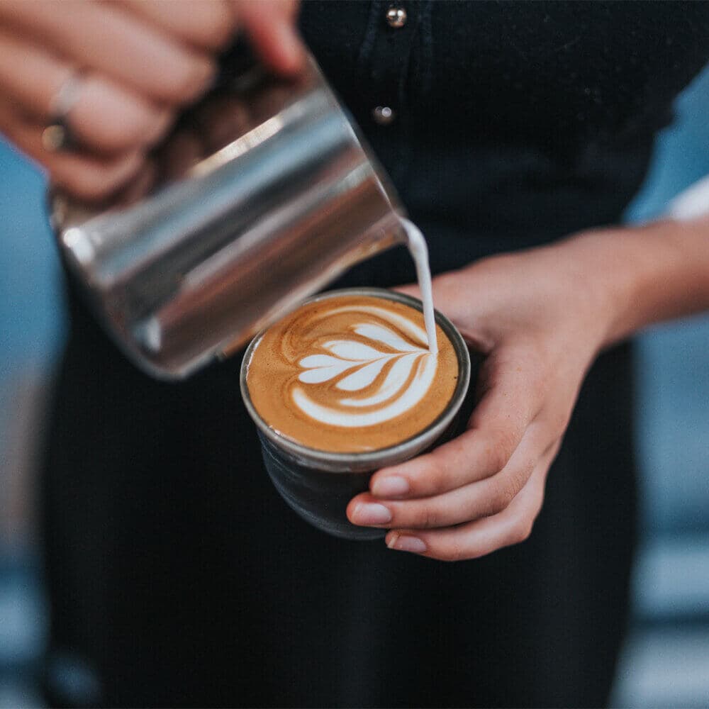 A person pouring milk from a metal jug into a coffee cup, creating latte art with a leaf design on top. - Home Instead