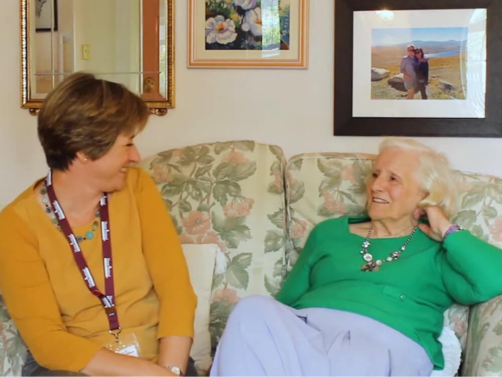 Two women sitting on a floral couch, smiling and talking in a cozy room with framed photos on the wall. - Home Instead