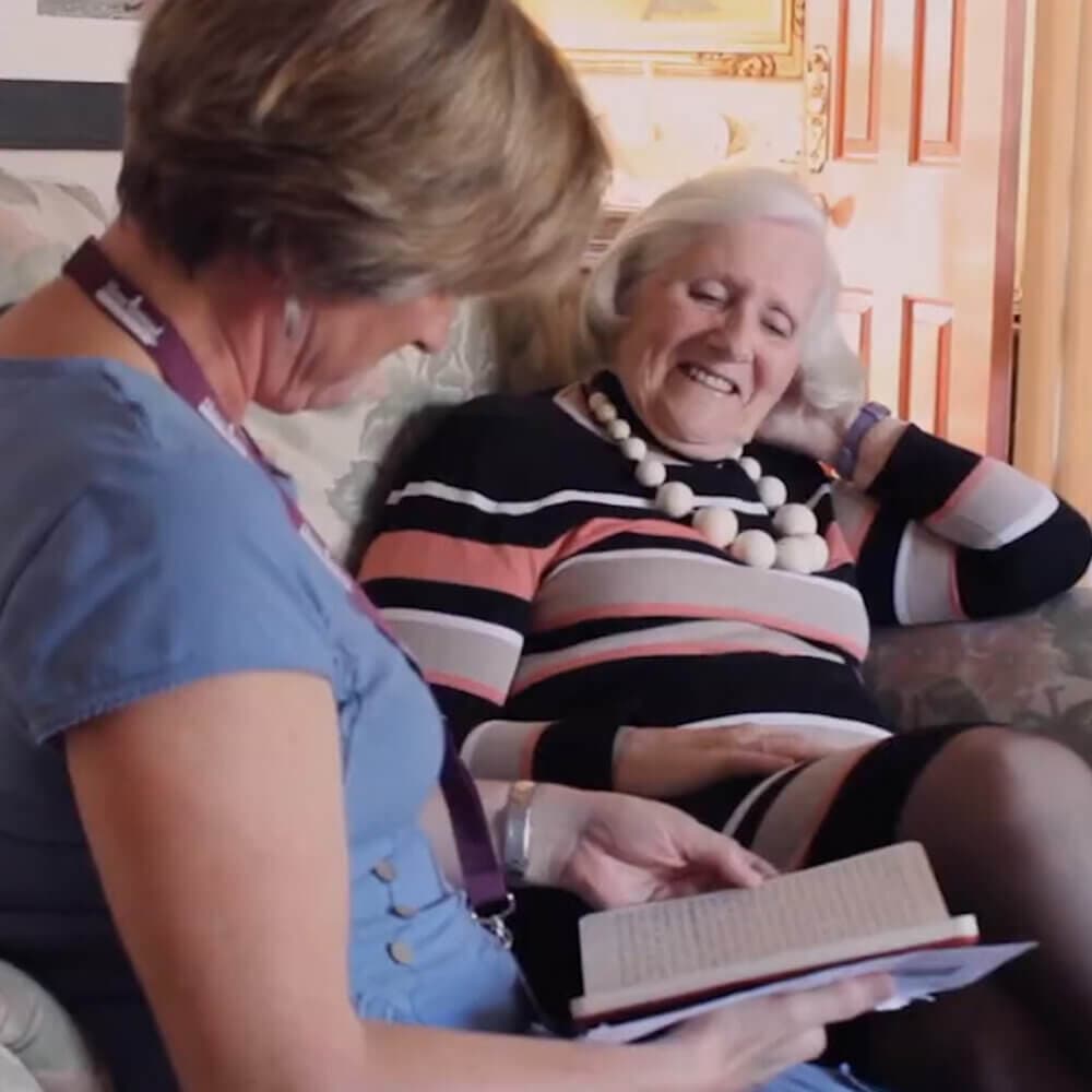 Two women sitting together on a couch, one reading a book aloud, both smiling and engaged in conversation. - Home Instead