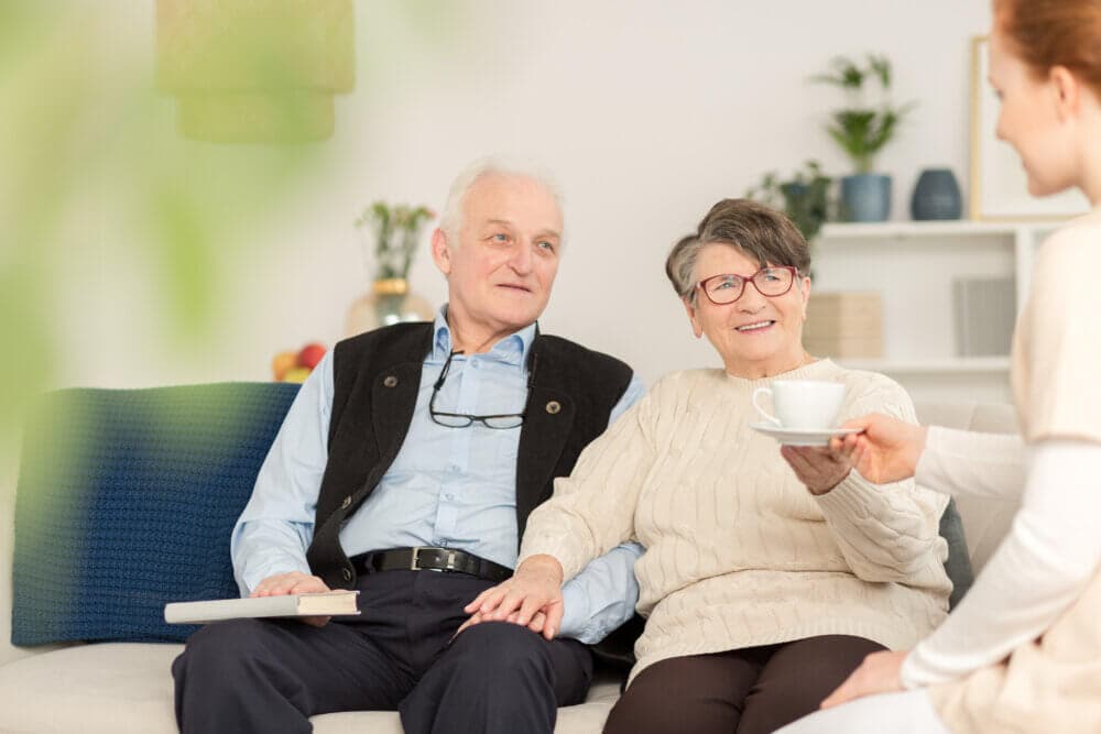 An elderly couple sits on a couch while a person hands them a cup of tea in a cozy living room setting. - Home Instead