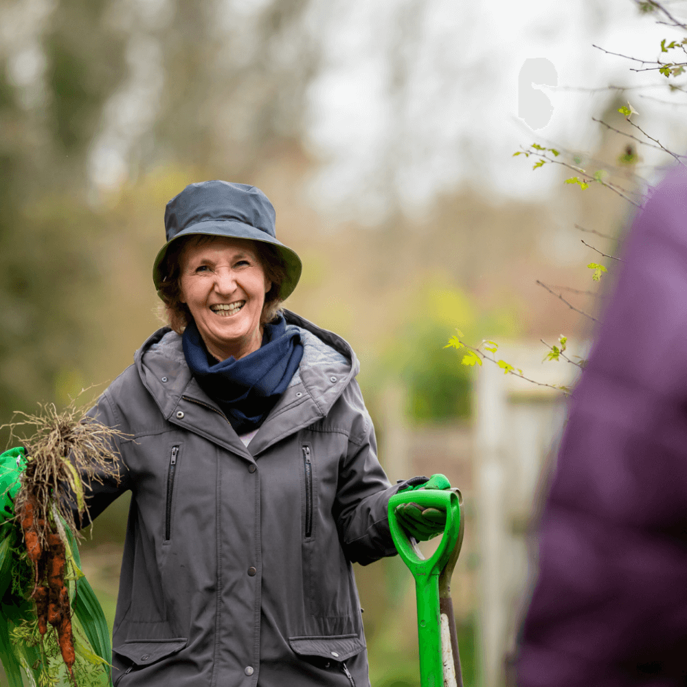 A smiling woman in a gray jacket and hat holds a bunch of freshly picked carrots and a green garden tool. - Home Instead