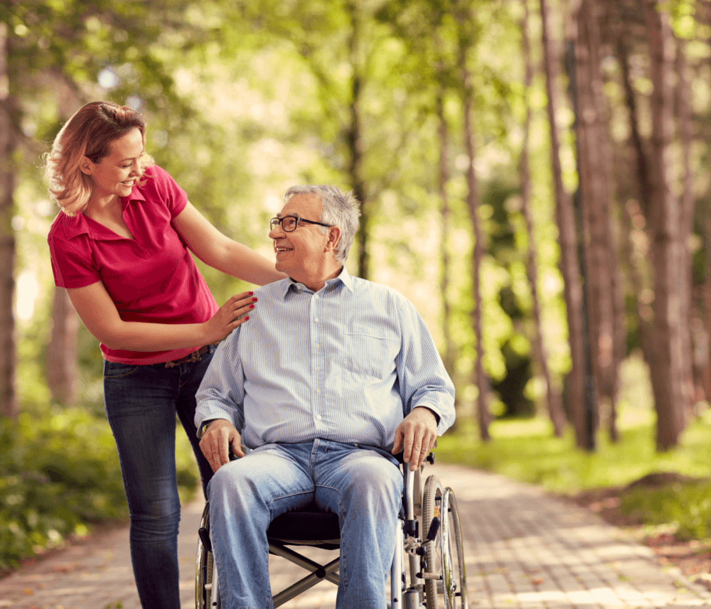 A smiling woman stands next to an elderly man in a wheelchair, sharing a moment in a sunlit, tree-lined park. - Home Instead