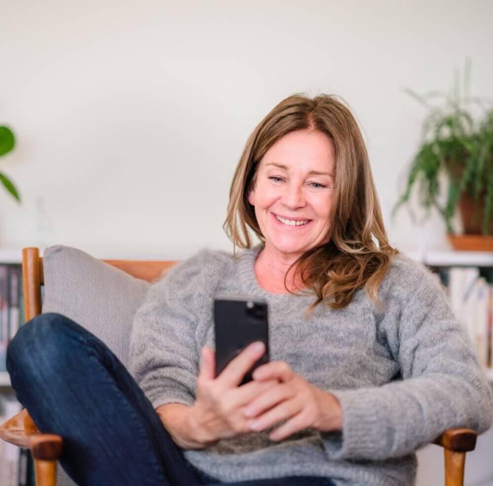 A woman with long hair smiles while looking at her phone, sitting on a chair with plants and bookshelves in the background. - Home Instead
