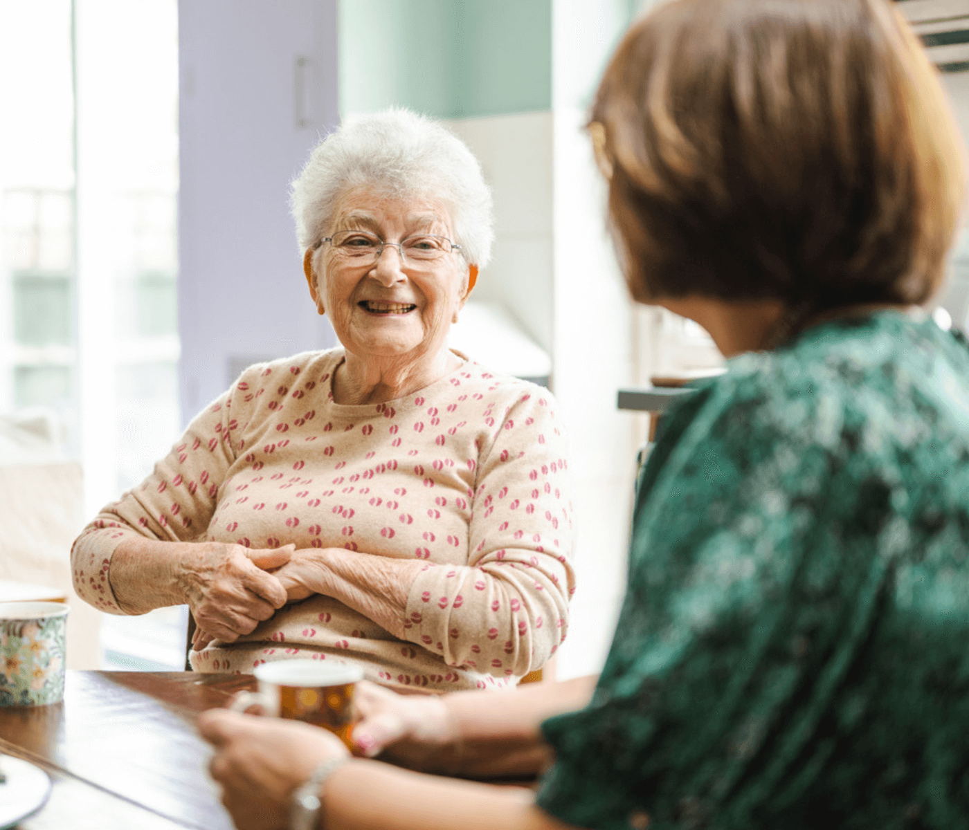 Elderly woman smiling and talking with another woman, both seated at a table with mugs. - Home Instead