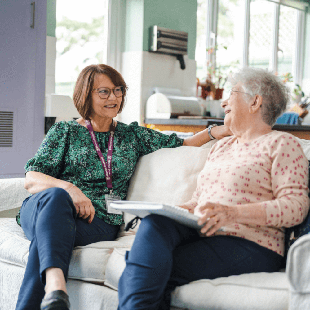 Two women sitting on a couch, smiling and chatting; one is holding a tablet, both appear to be engaged in conversation. - Home Instead