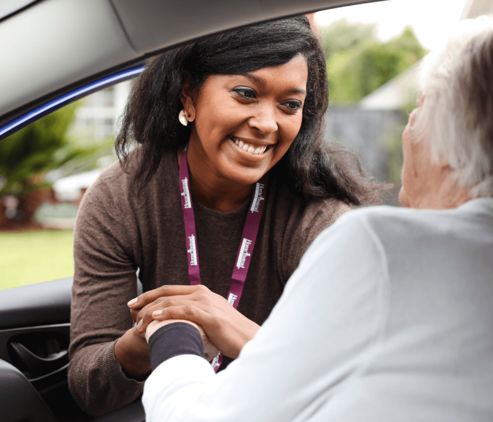 A woman smiling and holding the hand of a person seated in a car, offering reassurance. - Home Instead