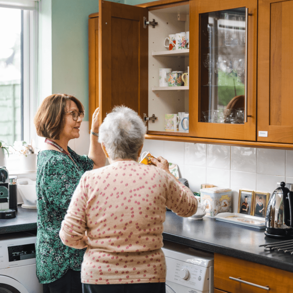 Two women talking in a kitchen with one reaching into a cabinet while the other gestures toward her. - Home Instead