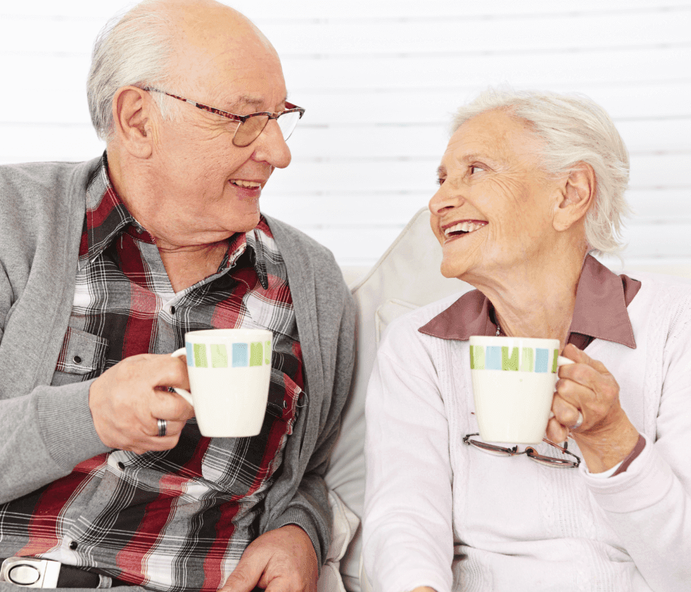 An elderly couple wearing sweaters, smiling at each other while holding mugs, sitting on a couch in a cozy setting. - Home Instead