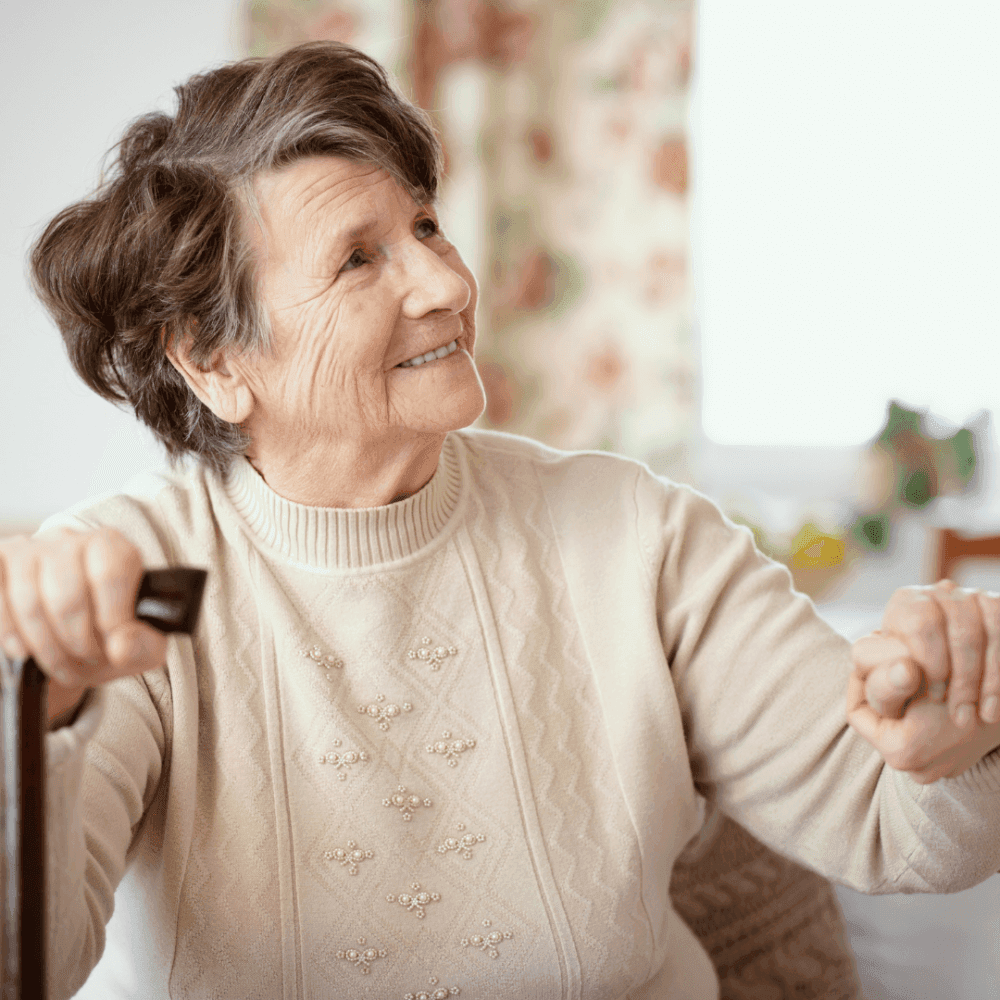 Elderly woman with short hair and a light sweater smiling while holding onto a cane. - Home Instead