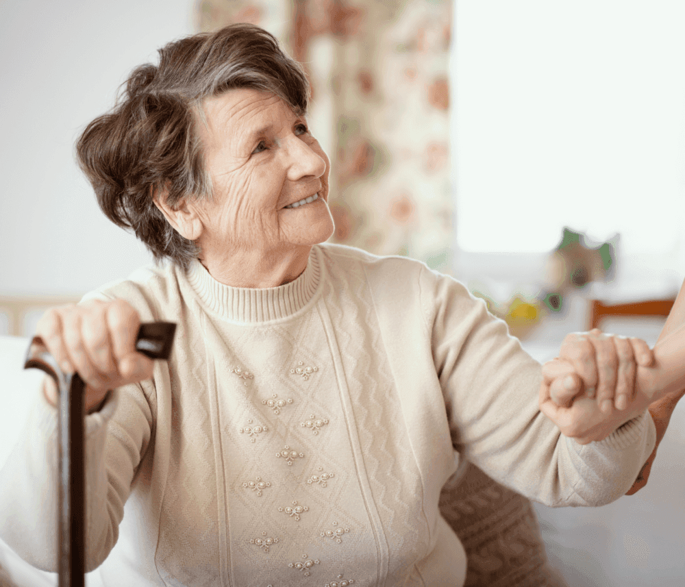 Elderly woman with a cane smiling and holding someone's hand, in a warmly lit room with floral curtains in the background. - Home Instead
