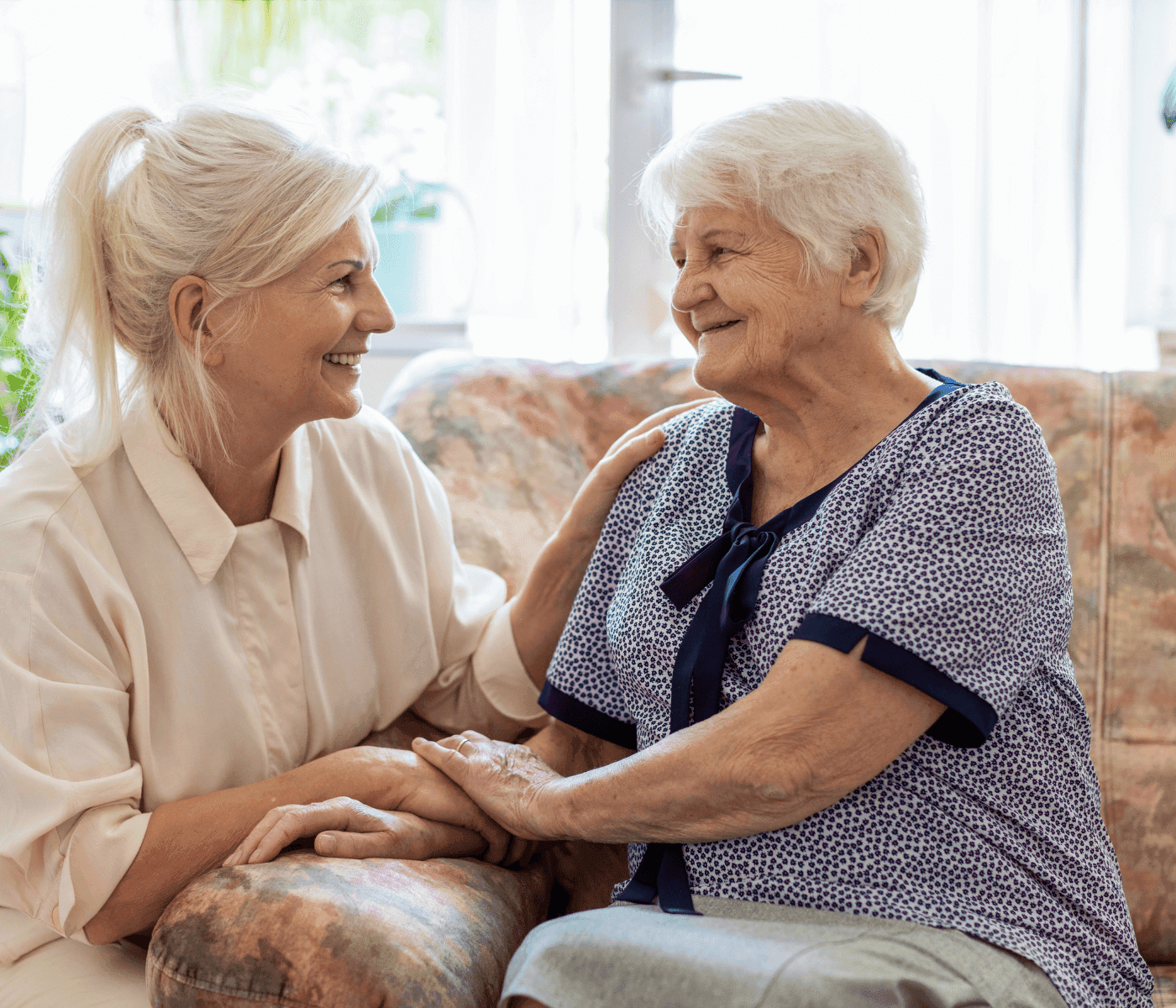 Two elderly women sitting on a couch, smiling and holding hands. - Home Instead
