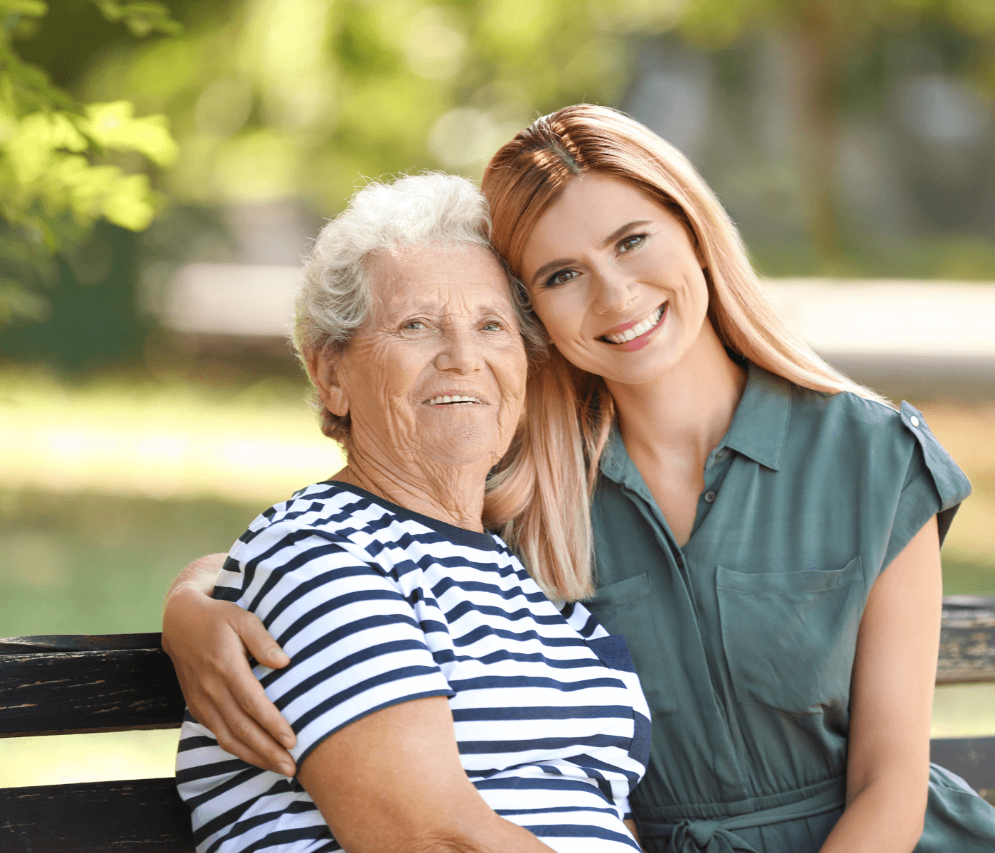 An elderly woman and a young woman sit on a bench outdoors, smiling and hugging each other. - Home Instead
