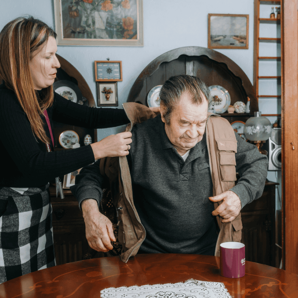 A woman helps an elderly man put on his jacket at a table with a purple mug on it. The room is decorated with paintings. - Home Instead