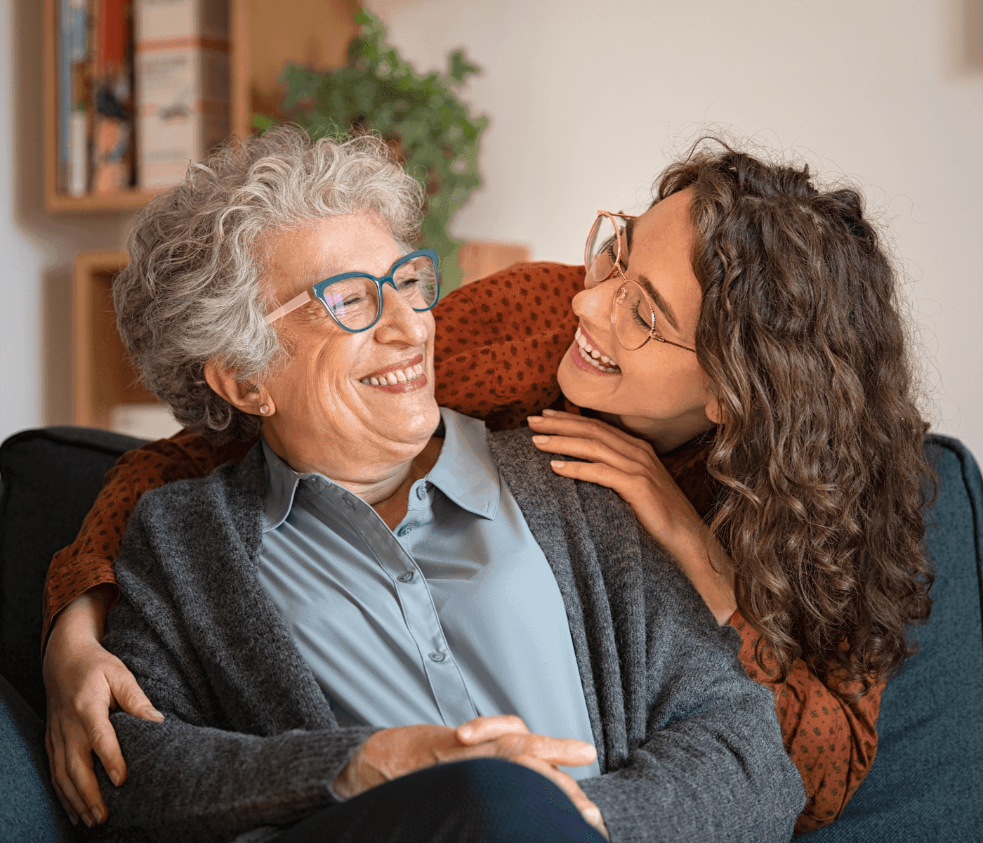 Two women wearing glasses share a joyful moment, smiling at each other while sitting on a sofa in a cozy room. - Home Instead