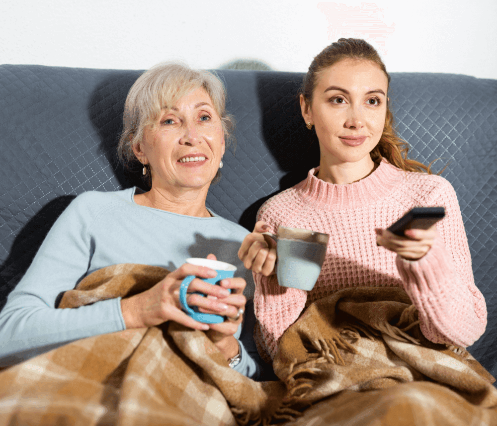 Two women sitting on a couch under a blanket, each holding a mug, while one woman points a remote at a TV. - Home Instead