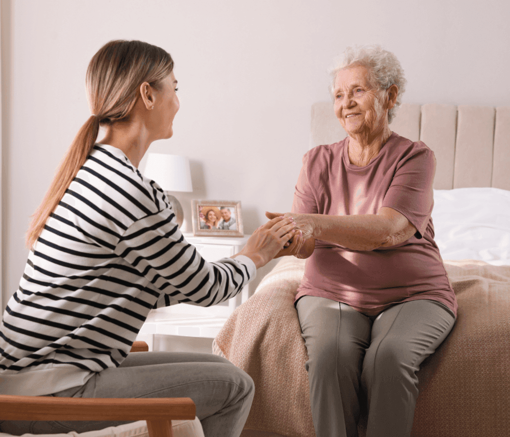 A young woman holds hands with an elderly woman sitting on a bed, both smiling warmly at each other. - Home Instead