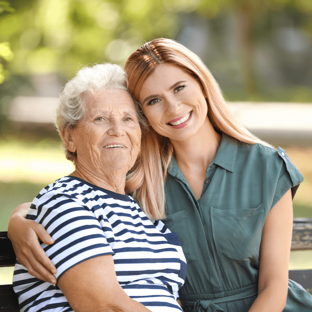 A young woman with long hair hugs an elderly woman in a striped shirt, both smiling outdoors on a sunny day. - Home Instead