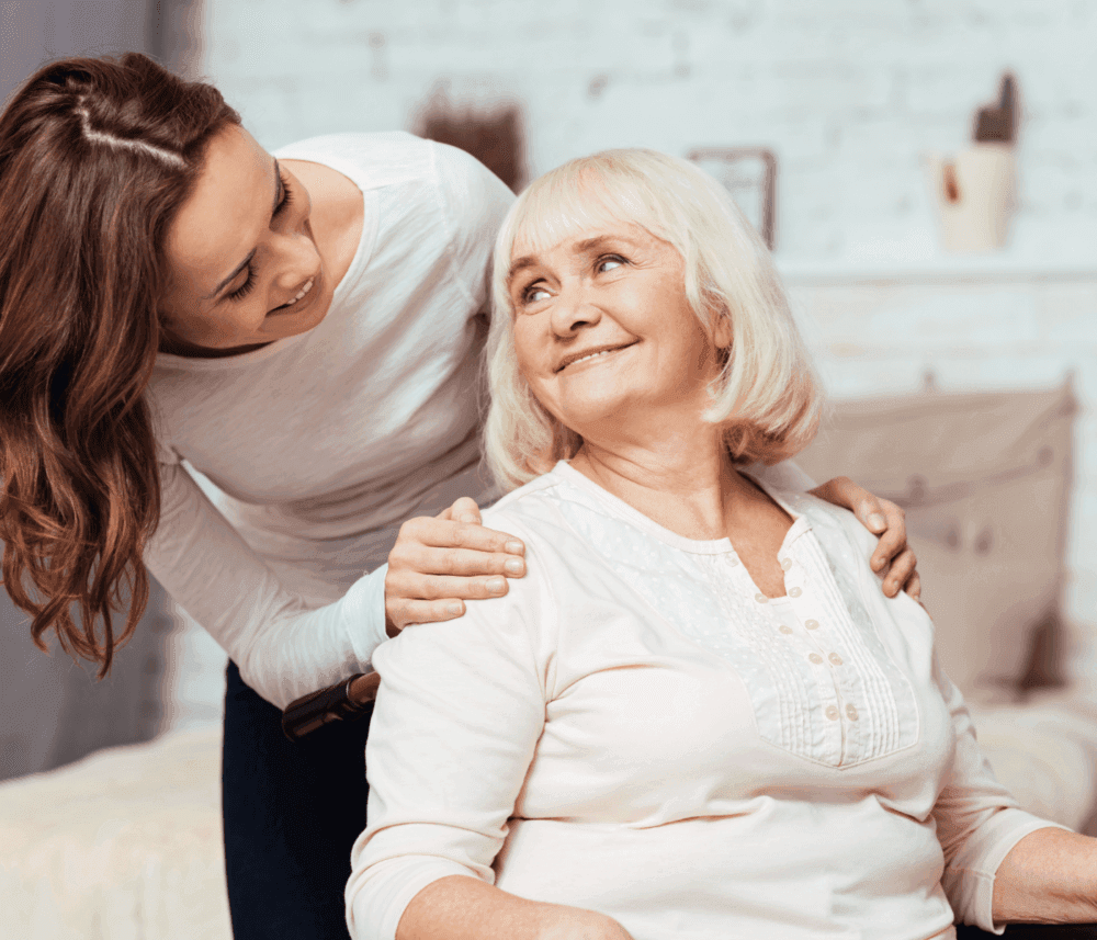 A smiling young woman stands behind an older woman, touching her shoulder, both sharing a joyful moment in a cozy setting. - Home Instead