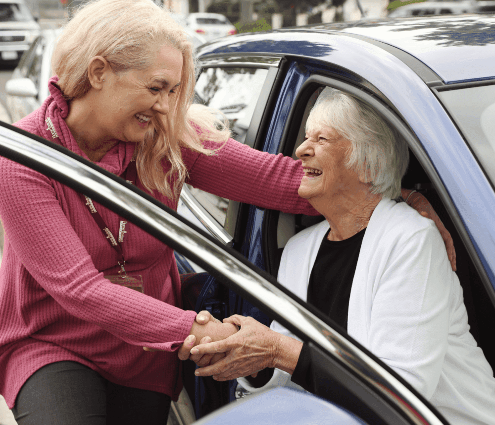 A woman helps an elderly woman out of a car; both are smiling and holding hands. - Home Instead
