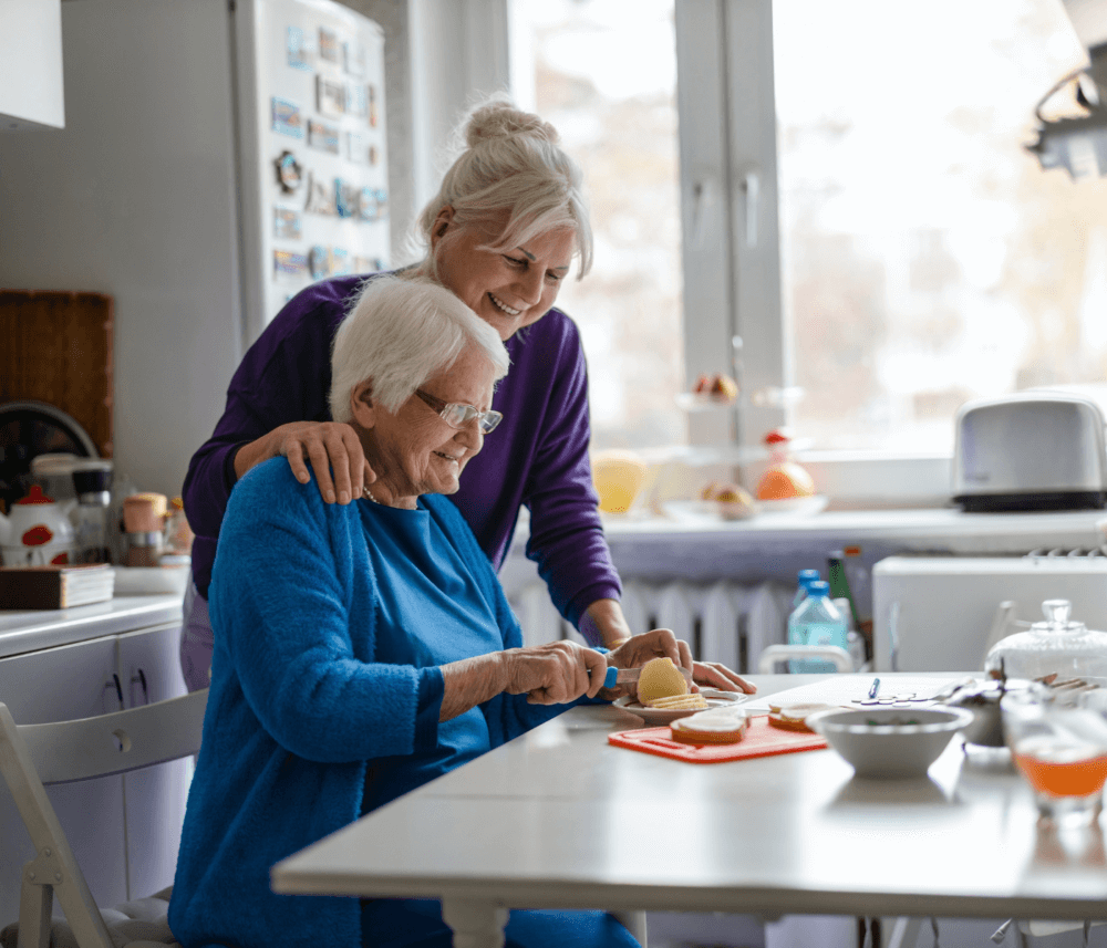 Two elderly women in a kitchen, one cutting food while the other stands behind her smiling supportively. - Home Instead