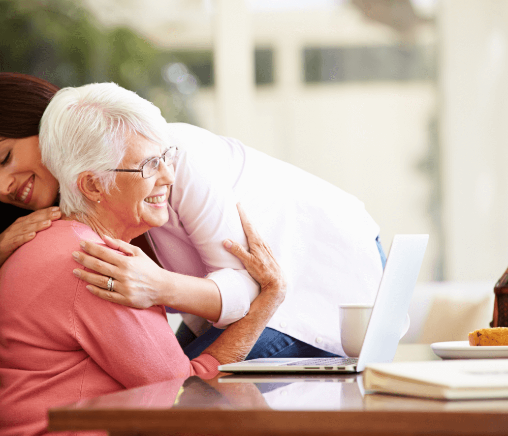 A younger woman hugs an older woman from behind as they sit at a table with a laptop, both smiling warmly. - Home Instead