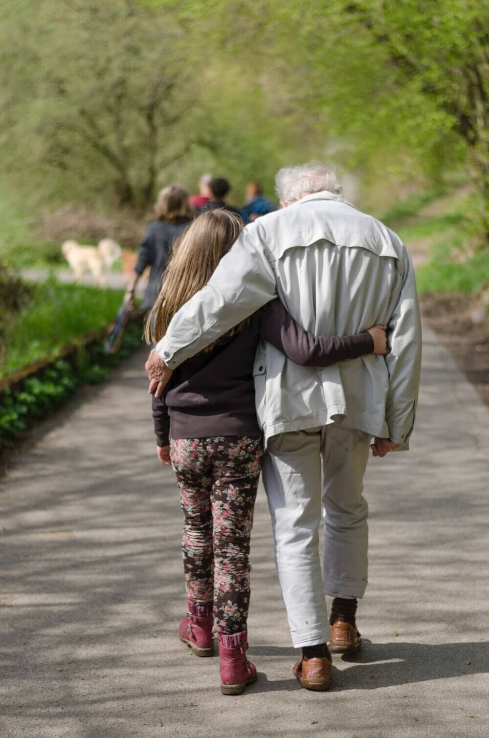 A young girl and elderly man walk arm-in-arm on a sunny path, surrounded by greenery. Their backs are to the camera. - Home Instead