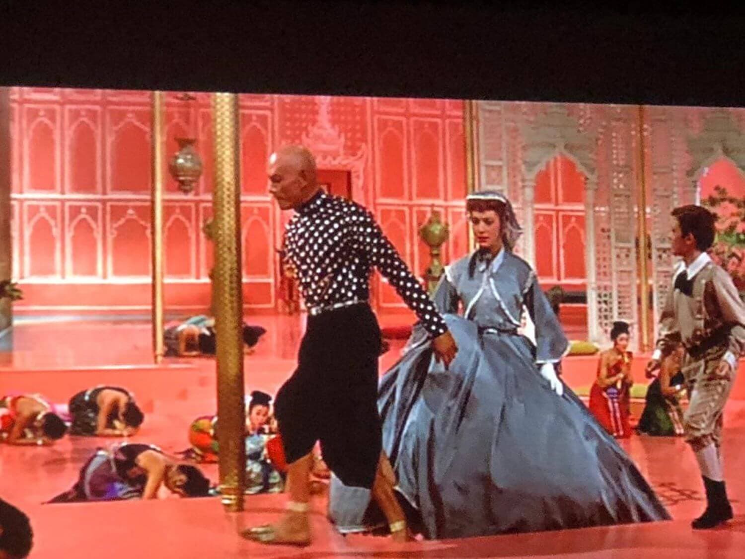 A bald man in a patterned shirt and a woman in a blue dress walk ahead of others bowing on a red floor. - Home Instead