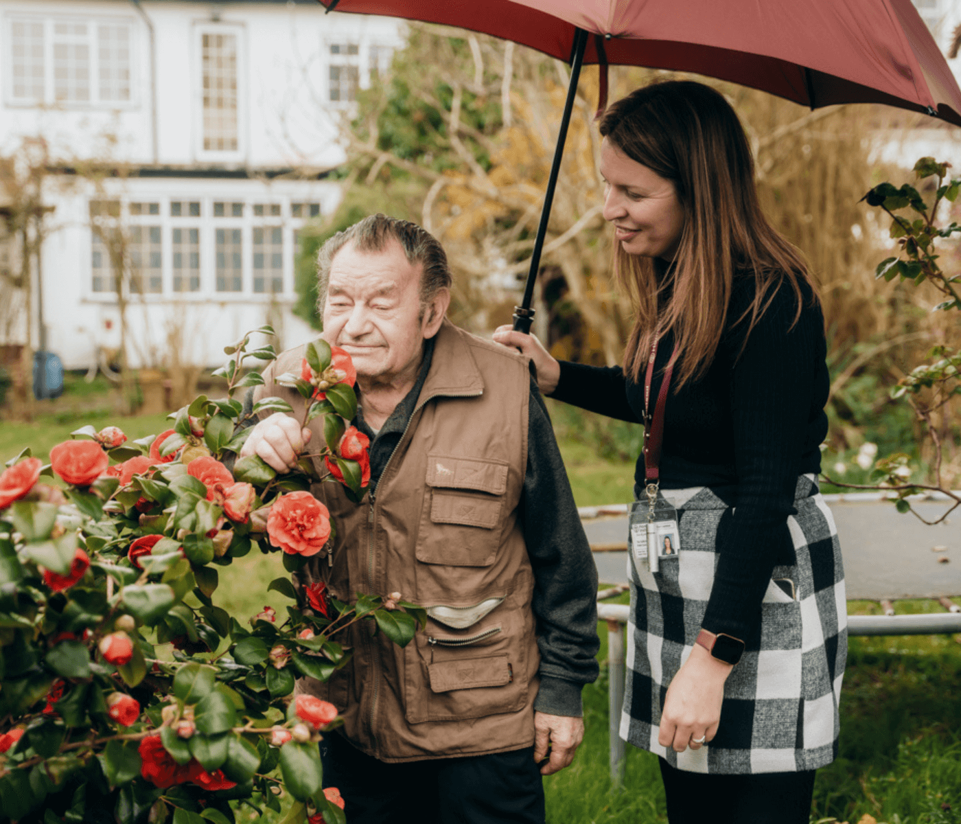 An elderly man and a woman holding a red umbrella stand by blooming red flowers in a garden. - Home Instead