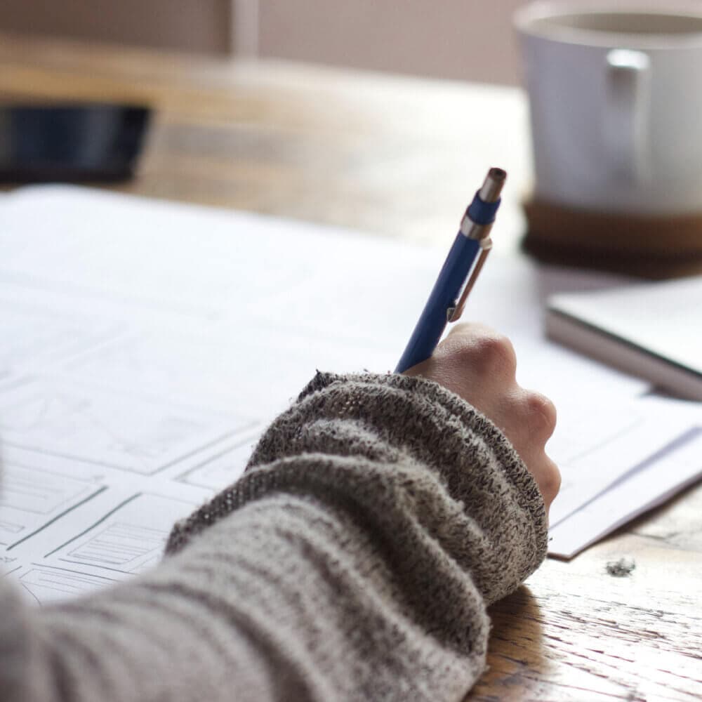 Person writing with a pen on a notebook at a wooden table, with a coffee cup and smartphone in the background. - Home Instead