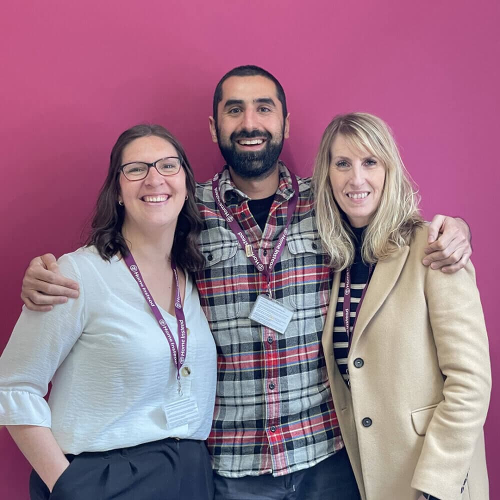 Three people standing together and smiling, with arms around each other, against a bright pink background. - Home Instead