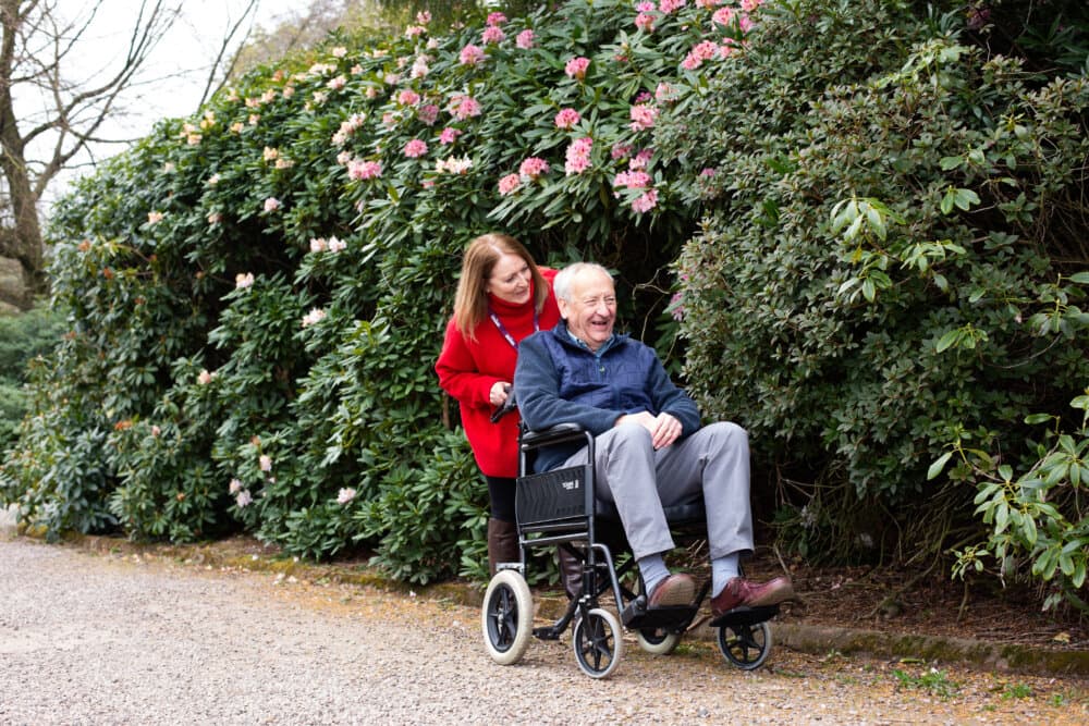 A woman in a red coat pushes an elderly man in a wheelchair along a path surrounded by lush green bushes and pink flowers. - Home Instead Bournemouth & Christchurch