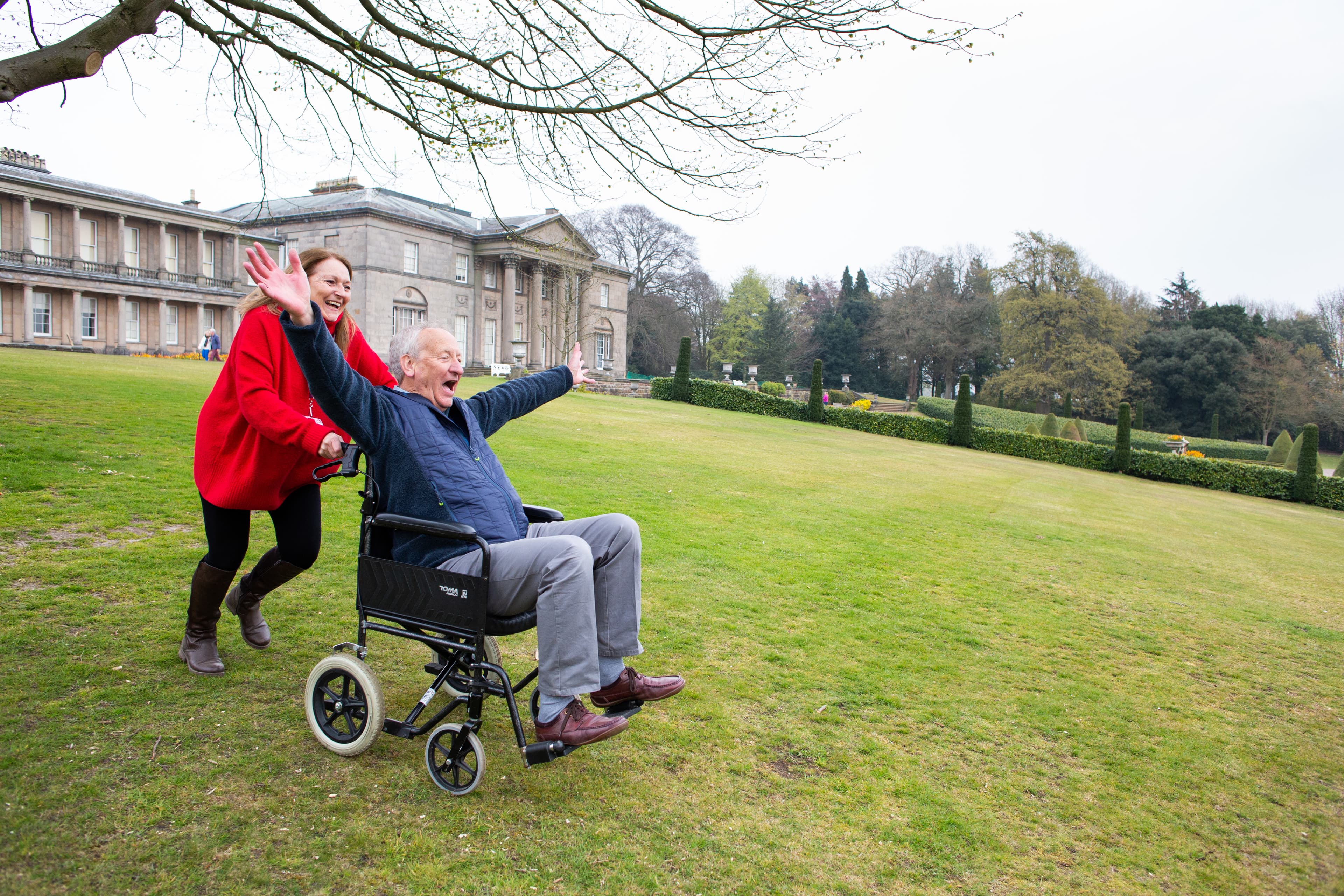 A woman joyfully pushes an elderly man in a wheelchair across a green lawn with a large, historic building in the background. - Home Instead