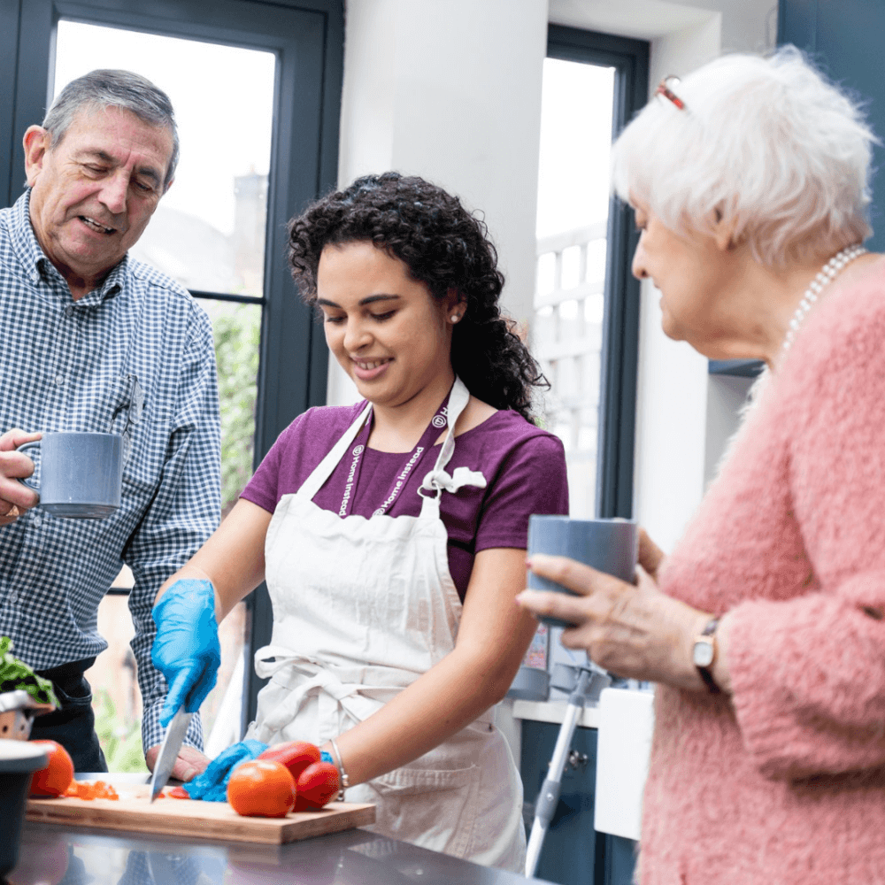 Young woman in an apron chopping vegetables in the kitchen, with two older adults holding mugs and smiling at her. - Home Instead